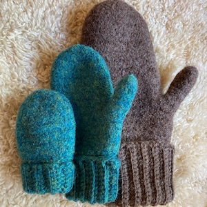 May include: A set of three wool mittens in varying sizes. One large mitten is dark brown with a ribbed cuff. Two smaller mittens are a vibrant teal blue with yellow flecks, also featuring ribbed cuffs. The texture is thick, felted, and fuzzy.