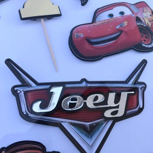 May include: A collection of birthday party decorations featuring a red race car, checkered flags, and a trophy. A large emblem with the name "Joey" in silver lettering is also included. The decorations are designed for a car-themed party.
