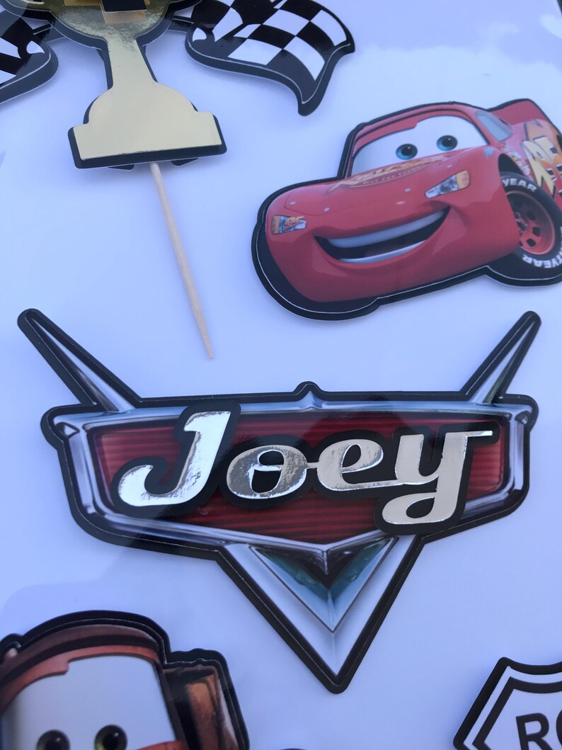 May include: A collection of birthday party decorations featuring a red race car, checkered flags, and a trophy. A large emblem with the name "Joey" in silver lettering is also included. The decorations are designed for a car-themed party.