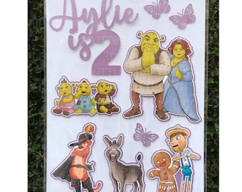 May include: A sheet of Shrek-themed birthday decorations. The design includes the name "Aylie is 2" in purple, along with illustrations of Shrek, Fiona, Donkey, Puss in Boots, and other characters. Butterflies are also included.