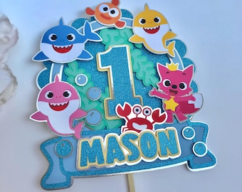 May include: A glittery blue cake topper featuring the number 1 and the name MASON in yellow letters. The design includes cartoon sharks, a crab, and an orange fish against a blue background with green seaweed and bubble accents.
