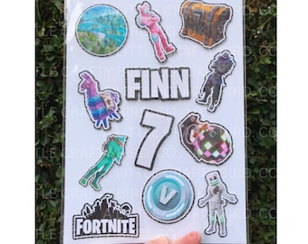 May include: A sheet of Fortnite-themed cake toppers featuring various game characters, a treasure chest, and the name "Finn" with the number "7". The Fortnite logo and a V-Bucks symbol are also included.