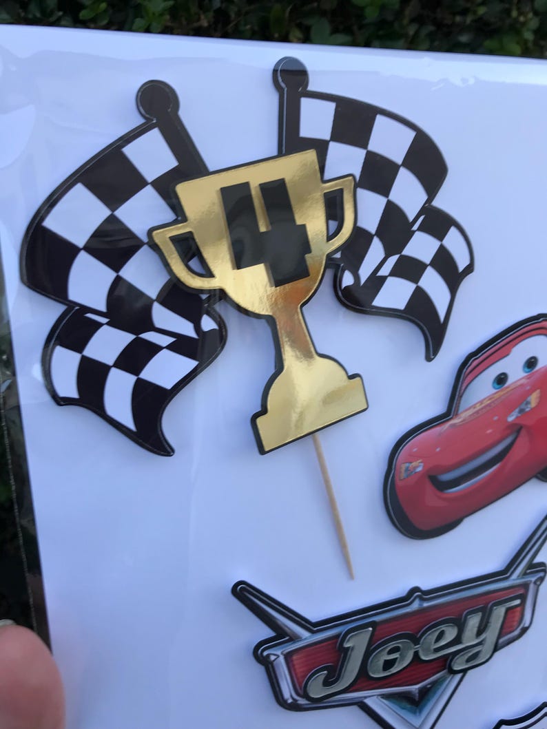 May include: Cake toppers featuring a gold trophy with the number 4, checkered flags, and a red race car. The word "Joey" is also visible in a racing-themed font. These decorations are perfect for a birthday party.