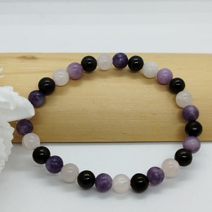 May include: A bracelet made of round beads in a variety of colours. The beads are black, light purple, pink, and white. The bracelet is presented on a light brown wooden surface.