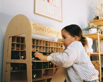 May include: A wooden toy garage with multiple compartments, labeled "Rumi's Garage", is filled with toy cars. A child is kneeling, interacting with the toy cars. A light blue wall and a framed picture are in the background.