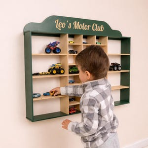 May include: A dark green wooden display shelf with multiple compartments, holding various toy cars. The shelf is labelled "Leo's Motor Club" in a cursive font. A child is reaching for a toy car on the shelf.
