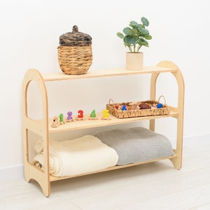 May include: A three-tiered wooden shelf unit with rounded sides. The top shelf holds a woven basket with a lid and a potted plant. The middle shelf displays a woven tray with toys and a wooden number train. The bottom shelf has folded blankets.