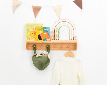 May include: A wooden wall shelf with pegs holds two children's books, Don't Tickle the Lion and Sunny Rainy, and a rainbow-shaped abacus. A green crocheted hanging basket with a small plush toy and a cream-colored sweater on a hanger are below.