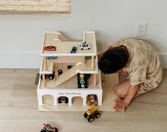 Puede incluir: Un garaje de estacionamiento de juguete de madera con múltiples niveles y rampas. El garaje es blanco con detalles de madera natural e incluye coches de juguete. El texto "My Parking" es visible en la parte delantera. Un niño está jugando con el juguete.