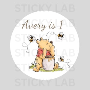 以下が含まれることがあります： 「Avery is 1」の文字と、ハチミツの壺の隣に座っているくまのプーさんのイラストが描かれた丸いステッカー。デザインには3匹のハチが含まれており、白い背景に設定されています。