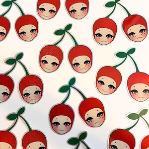 May include: A set of 14 red and green glitter cherry stickers with cartoon faces. Each sticker features a cherry with a stem and leaf, and a face with eyes, eyelashes, and a mouth.