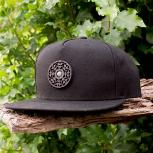 Peut inclure: Une casquette de baseball noire avec un motif rond en bois découpé au laser, orné d'une pierre précieuse verte au centre.
