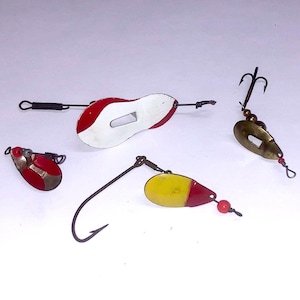May include: Four fishing lures with different colors and designs. The lures are made of metal and have hooks. One lure is white with red accents, another is yellow with red accents, another is red with white accents, and the last one is gold with a black hook.
