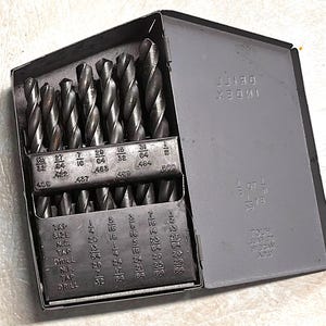 May include: A set of drill bits in a gray metal case. The case is open, revealing multiple drill bits of varying sizes. The case has the text "INDEX DRILL BITS" and size markings. A useful tool for woodworking and metalworking projects.