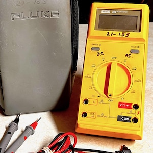May include: A yellow Fluke multimeter with a black and gray carrying case. The multimeter has a digital display and various settings. Included are test leads and a black and red cable. The case is gray and has the text "21-153" and "Fluke".