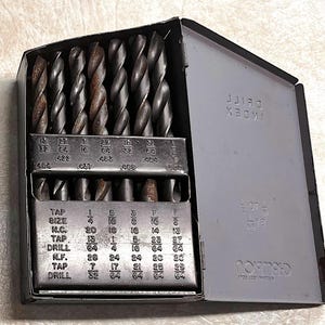 May include: A gray metal drill bit index case, open to reveal a set of drill bits. The case has the word "INDEX" printed on it. The drill bits are arranged in rows and columns, with size markings.