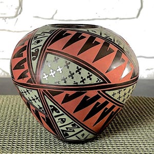 Może przedstawiać: Okrągły, ręcznie malowany ceramiczny wazon o unikalnym geometrycznym wzorze. Wazon ma wzór czerwonych, czarnych i jasnozielonych kształtów. Wzór zawiera trójkąty, zygzaki i symbole w kształcie krzyża. Wazon stoi na teksturowanej powierzchni.