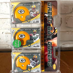 May include: A clear plastic display case holding three Green Bay Packers mini helmet cards. Each card features a yellow helmet with a green "G" logo. The display also includes the text "Green Bay Gamers" and player names.