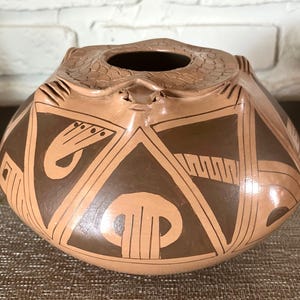 Puede incluir: Un jarrón de cerámica artesanal de color beige, con un cuerpo redondeado y una pequeña abertura en la parte superior. El jarrón está adornado con diseños geométricos y estilizados en un tono marrón más oscuro, creando un contraste llamativo.