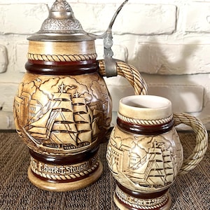 May include: Two ceramic beer steins with a nautical theme. The larger stein has a metal lid and handle. Both steins are tan with brown accents and feature a detailed relief of a sailing ship and the text "Bark T. Staysail".