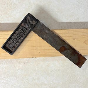May include: A vintage metal try square tool with a dark, textured handle featuring the embossed text SOUTHINGTON. The long, flat blade shows signs of oxidation and wear, with a dark grey finish and a distinct rust spot near the end of the metal.