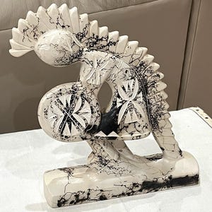 Puede incluir: Escultura decorativa en blanco y negro de un caballo estilizado. La escultura tiene una superficie texturizada con vetas negras. El caballo sostiene un objeto redondo con un diseño de cruz. La escultura está montada sobre una base rectangular.