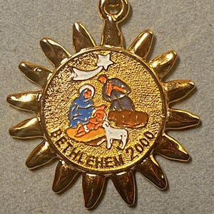 Pode incluir: Pingente dourado com design de raios solares. O centro apresenta uma imagem circular da cena da Natividade com o texto "BETHLEHEM 2000". O pingente tem uma argola para corrente.