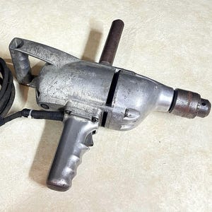 May include: A heavy-duty silver metal electric drill featuring a top-mounted handle, a pistol-grip handle with a trigger, and a black power cord. The tool has a weathered, scratched finish on the main housing and a dark metal chuck at the front.