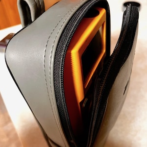 Op de afbeelding: Een grijze leren etui met een zwarte ritssluiting, gedeeltelijk open om een oranje voorwerp binnenin te onthullen. De etui heeft een handvat en ligt op een lichtgekleurd oppervlak.