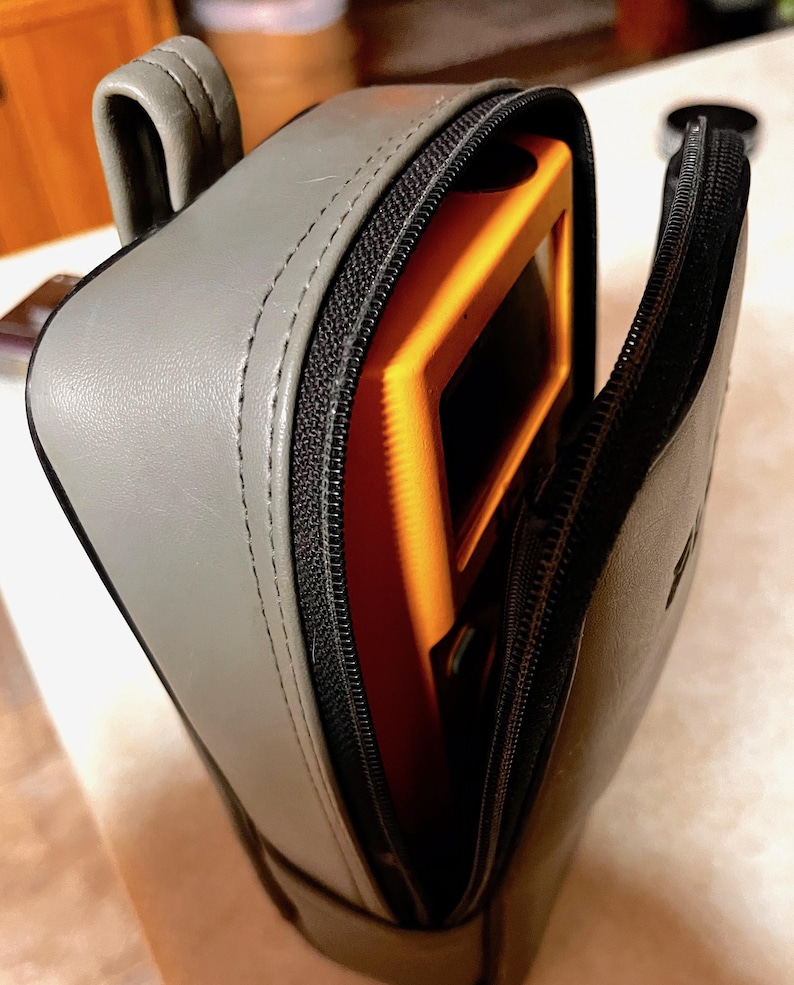 Op de afbeelding: Een grijze leren etui met een zwarte ritssluiting, gedeeltelijk open om een oranje voorwerp binnenin te onthullen. De etui heeft een handvat en ligt op een lichtgekleurd oppervlak.