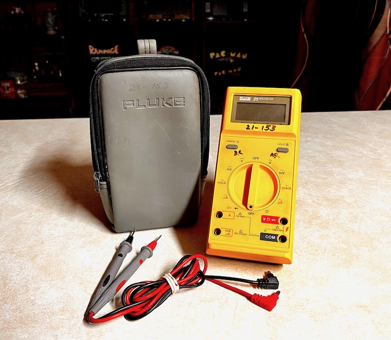 May include: A yellow Fluke multimeter with a black and gray carrying case. The multimeter has a digital display and various settings. Included are test leads and a black and red cable. The case is gray and has the text "21-153" and "Fluke".