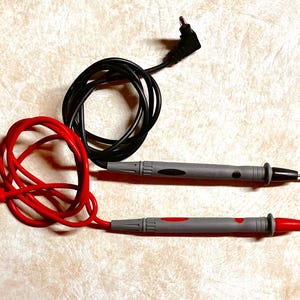 Op de afbeelding: Twee elektrische testkabels met grijze handgrepen en zwarte en rode accenten. E&eacute;n kabel heeft een rode kabel en de andere een zwarte kabel. De kabels worden gebruikt voor het testen van elektrische circuits en componenten.