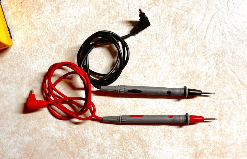 Op de afbeelding: Twee elektrische testkabels met grijze handgrepen en zwarte en rode accenten. E&eacute;n kabel heeft een rode kabel en de andere een zwarte kabel. De kabels worden gebruikt voor het testen van elektrische circuits en componenten.