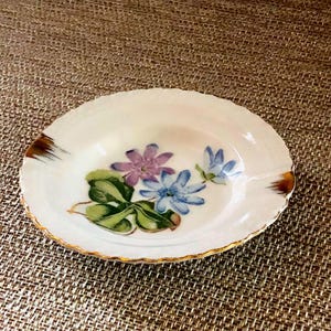 以下が含まれることがあります： 楕円形のオフホワイトのセラミック皿、花柄のデザイン。 金色の縁とアクセントが特徴です。 花柄のデザインには、紫と青の花と緑の葉が含まれています。 この皿は、装飾またはサービングを目的としている可能性があります。