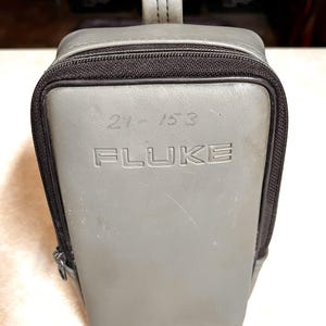 Op de afbeelding: Een grijze Fluke draagtas met een zwarte rits en handvat. Het woord "FLUKE" is in reli&euml;f op de voorkant aangebracht, samen met de nummers "21-153". De tas is rechthoekig met afgeronde hoeken.