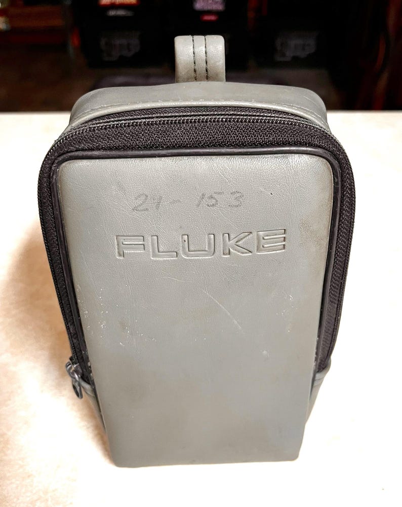 Op de afbeelding: Een grijze Fluke draagtas met een zwarte rits en handvat. Het woord "FLUKE" is in reli&euml;f op de voorkant aangebracht, samen met de nummers "21-153". De tas is rechthoekig met afgeronde hoeken.