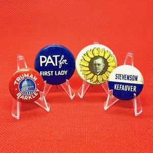 Puede incluir: Cuatro botones de campaña política vintage sobre soportes de plástico transparente. Los botones presentan texto e imágenes, incluyendo "Truman Barkley", "Pat for First Lady", un retrato dentro de un girasol y "Stevenson Kefauver". El fondo es rojo liso.