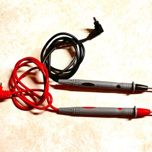 Op de afbeelding: Een paar grijze multimeter testkabels met zwarte en rode draden. De kabels hebben scherpe metalen punten voor elektrisch testen. De rode kabel is verbonden met een rode connector, de zwarte met een zwarte connector.