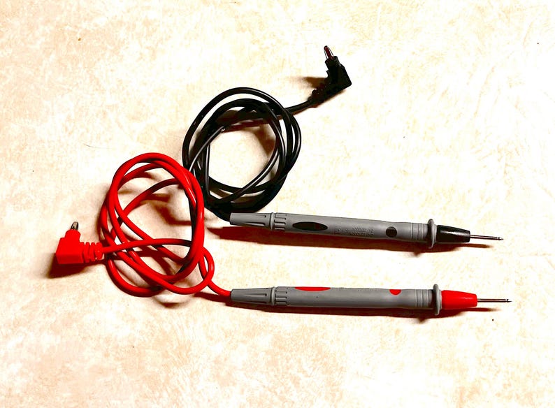 Op de afbeelding: Een paar grijze multimeter testkabels met zwarte en rode draden. De kabels hebben scherpe metalen punten voor elektrisch testen. De rode kabel is verbonden met een rode connector, de zwarte met een zwarte connector.