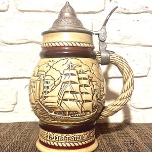 May include: A beige ceramic beer stein with a detailed relief of a sailing ship and buildings. It has a metal lid and handle, and a rope-like handle. The base has the text "The Bark & Staysail Still".