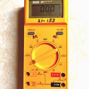 Op de afbeelding: Een gele Fluke 25 multimeter met een display dat "00.0" weergeeft. Het apparaat heeft instellingen voor spanning, stroom en weerstand, met labels zoals "D.C.", "A" en "COM".