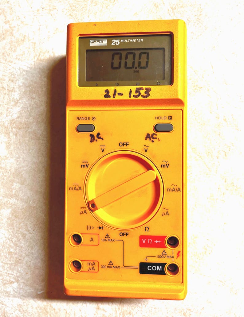 Op de afbeelding: Een gele Fluke 25 multimeter met een display dat "00.0" weergeeft. Het apparaat heeft instellingen voor spanning, stroom en weerstand, met labels zoals "D.C.", "A" en "COM".