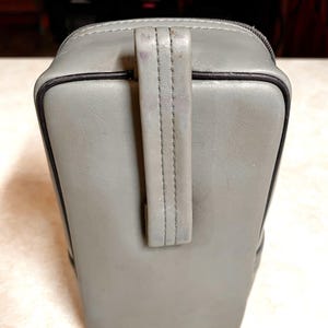 Op de afbeelding: Een grijze rechthoekige tas met een handvat. De tas heeft een ritssluiting en zwarte rand. Het handvat is aan de bovenkant van de tas gestikt. De tas lijkt gemaakt van een leerachtig materiaal.