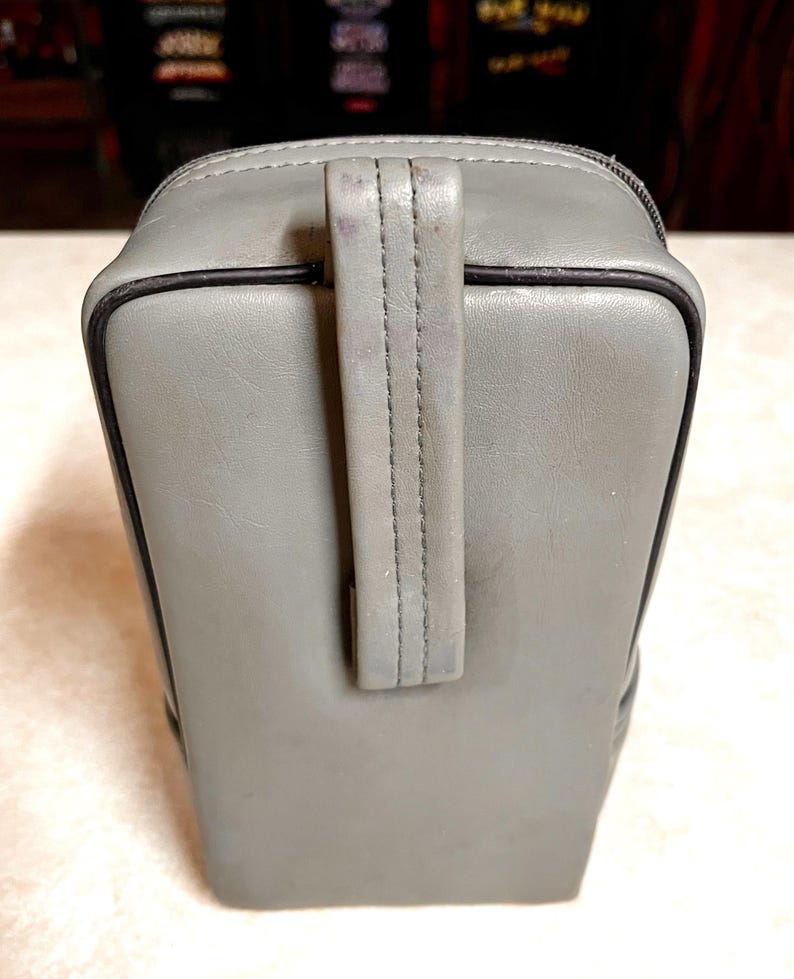 Op de afbeelding: Een grijze rechthoekige tas met een handvat. De tas heeft een ritssluiting en zwarte rand. Het handvat is aan de bovenkant van de tas gestikt. De tas lijkt gemaakt van een leerachtig materiaal.