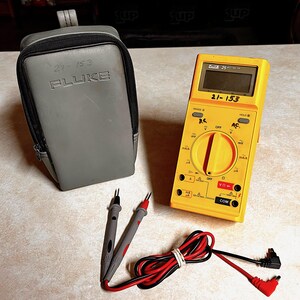 Op de afbeelding: Een gele Fluke 25 multimeter met een grijze draagtas en testkabels. De multimeter heeft een digitaal display en diverse instellingen. De tas is grijs met de tekst "21-153 FLuke". De testkabels zijn grijs, rood en zwart.