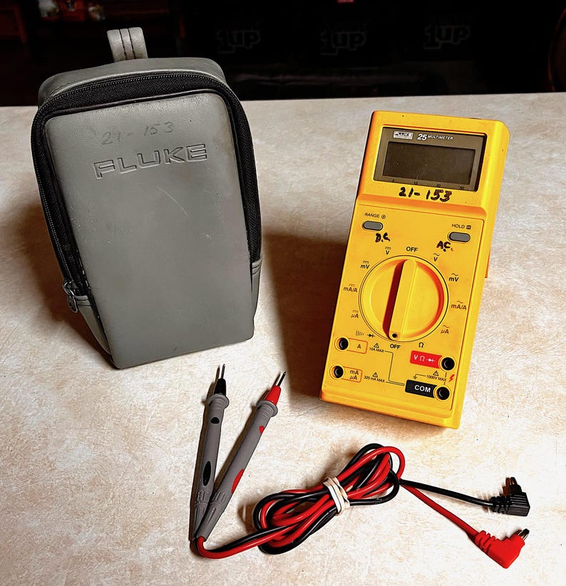 Op de afbeelding: Een gele Fluke 25 multimeter met een grijze draagtas en testkabels. De multimeter heeft een digitaal display en diverse instellingen. De tas is grijs met de tekst "21-153 FLuke". De testkabels zijn grijs, rood en zwart.