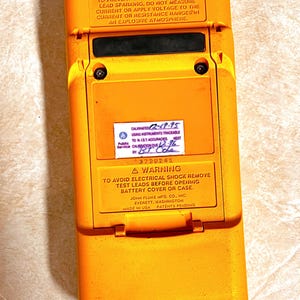 Op de afbeelding: Een gele en zwarte Fluke multimeter met tekstwaarschuwingen en kalibratie-informatie. Het apparaat heeft een rechthoekige vorm met een scharnierend compartiment. De tekst bevat waarschuwingen over explosie en elektrische schokken.