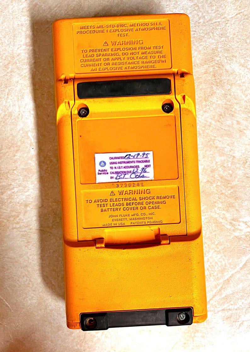Op de afbeelding: Een gele en zwarte Fluke multimeter met tekstwaarschuwingen en kalibratie-informatie. Het apparaat heeft een rechthoekige vorm met een scharnierend compartiment. De tekst bevat waarschuwingen over explosie en elektrische schokken.