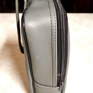 Op de afbeelding: Een grijze, rechthoekige etui met een zwarte ritssluiting en een gebogen handvat. De etui lijkt gemaakt van een glad, leerachtig materiaal. De ritssluiting loopt langs de zijkant van de etui.
