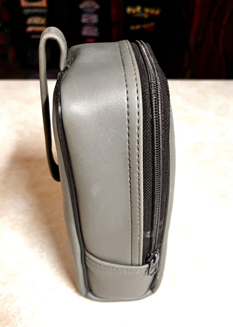 Op de afbeelding: Een grijze, rechthoekige etui met een zwarte ritssluiting en een gebogen handvat. De etui lijkt gemaakt van een glad, leerachtig materiaal. De ritssluiting loopt langs de zijkant van de etui.